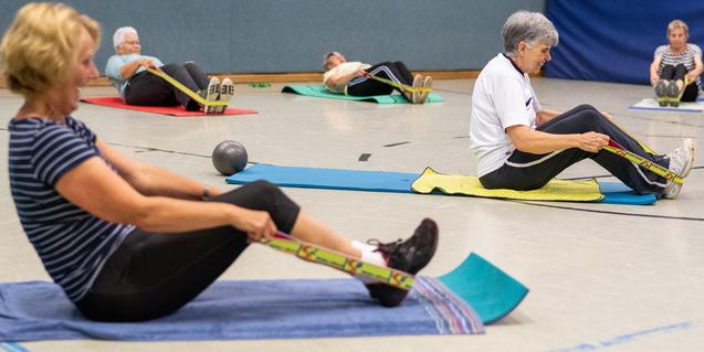 Senioren führen Rückenübungen mit Widerstandsbändern auf Matten in einer Sporthalle durch. Diese sind in verschiedenen Farben.