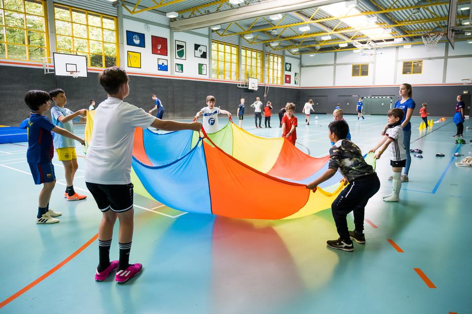 Gruppe von Kindern in einer Sporthalle, die einen bunten Parachute in die Höhe halten und gemeinsam spielen.