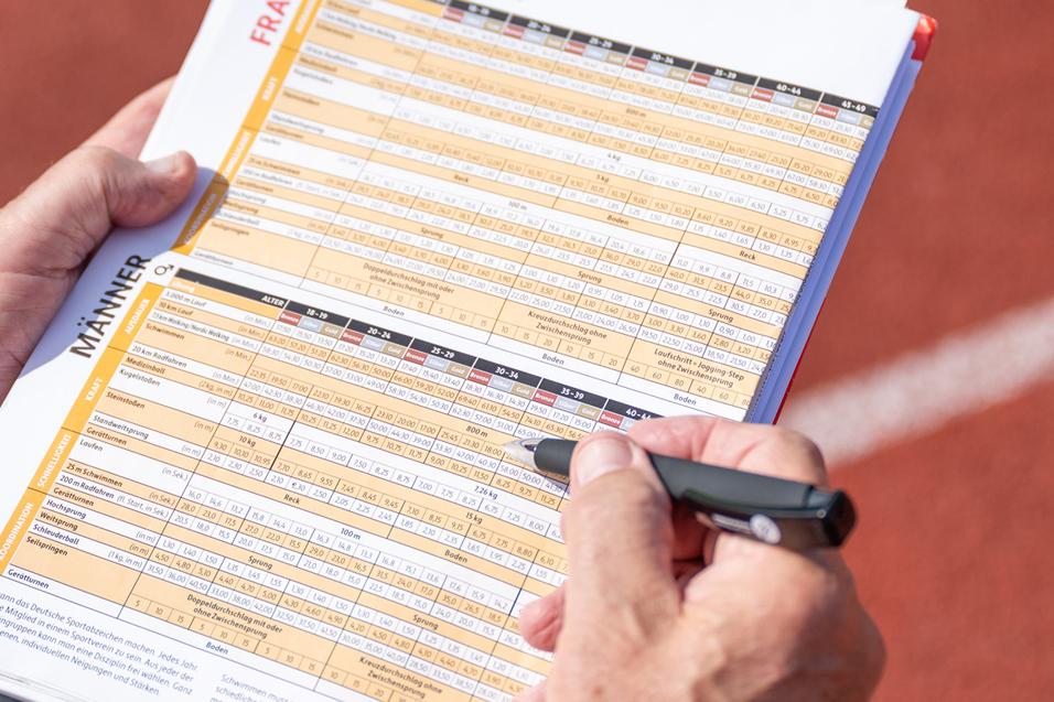 Hand eines Prüfers, der auf einem Blatt mit Sportabzeichen-Anforderungen auf einer Laufbahn notiert.