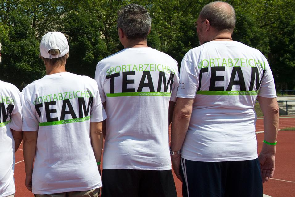 Gruppe von sieben Personen in weißen T-Shirts mit dem Aufdruck "TEAM" auf einer Laufbahn, Blick von hinten.