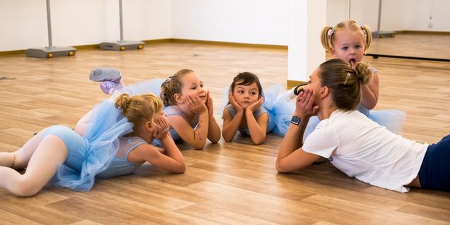 Fünf kleine Balletttänzerinnen in blauen Tüllkleidern liegen lächelnd auf dem Boden und betrachten eine Trainerin.