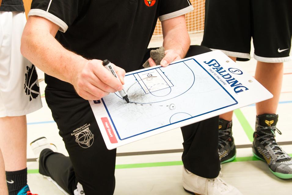 Trainer kniet mit Taktiktafel und zeichnet Spielzüge, während Spieler im Hintergrund stehen und einen Basketball halten.