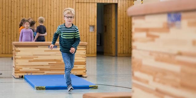 Ein Junge in gestreiftem Pullover läuft über eine Übungsfläche, während Erwachsene und Kinder im Hintergrund aktiv sind.